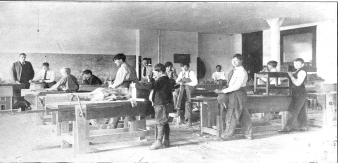 An image of a shop room at CWU in 1908 from the 1908-09 CWU catalogue. There are several students in the room standing next to a work desk. There are several desks in the room and an instructor standing at the front of the room.